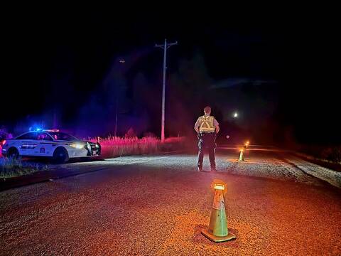 A BC Highway Patrol officer mans an impaired driving check stop on a lonely side road at night using lighted cones and his police cruiser