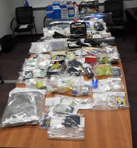 A large number of items are displayed in clear evidence bags on a table, including firearms, cash, pills, drugs, brass knuckles and contraband tobacco.