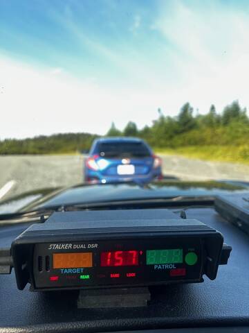 A blue car is pulled over on the side of a road. A police vehicle’s radar is pictured in the foreground displaying a speed of 157 km/h.
