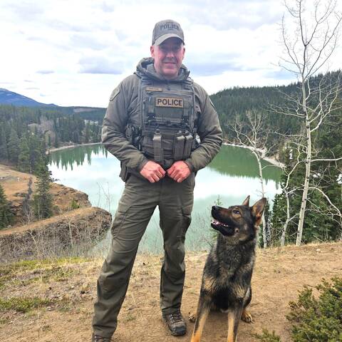 Corporal Cam Long stands with Police Service Dog Riggs on a scenic overlook above a river and forest.