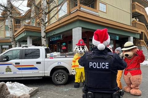 A photo, from the 2024 Whistler Emergency Services Holiday Food and Toy Drive, of a Royal Canadian Mounted Police truck with Fire Officer Sparky and RCMP Safety Bear wearing a red serge standing at the back of the police truck, and the back of a RCMP officer wearing a red Santa hat