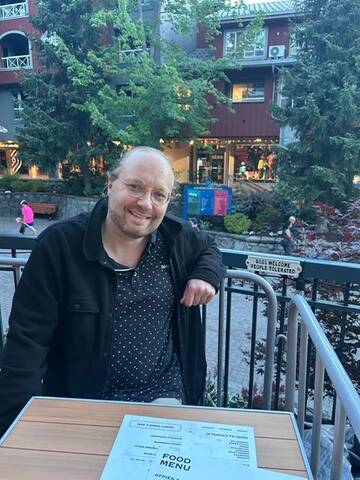 Man with blond hair, round glasses and a black jacket sitting at a table.