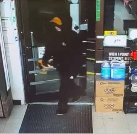 An individual steps inside the store. The person is wearing a black coat, a pair of black pants, a pair of black and white shoes, a black hoodie, a pair of black and red work gloves and a yellow/orange base ball cap;