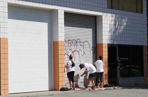 Trois personnes s’affairent à effacer des graffitis sur une porte coulissante en tôle ondulée d’un immeuble commercial.
